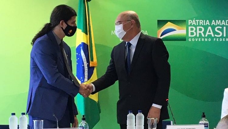 A imagem mostra dois homens, apertando as mãos. Atrás o banner verde e amarelo com os dizeres 