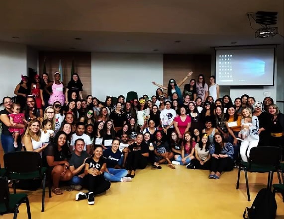 Grupo de homens e mulheres, estudantes de Enfermagem, sentados no chão. Alguns usam adereços e fantasias, na confraternização final do evento.