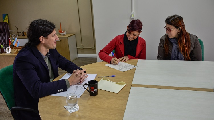A imagem mostra três pessoas sentadas à mesa, assinado papéis.