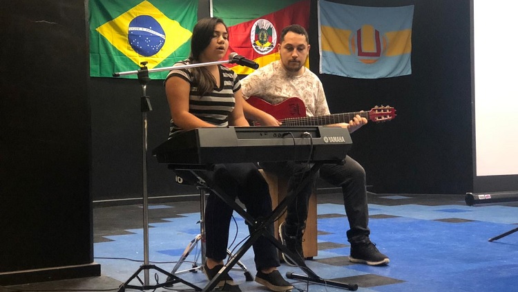 Uma menina canta enquanto toca teclado. Ao lado dela, um homem toca violão. Eles estão em um palco. Ao fundo, aparecem as bandeiras do Brasil, do Rio Grande do Sul e da FURG. 