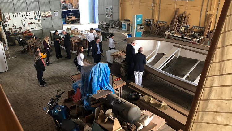 A imagem mostra o ambiente de uma oficina de trabalho, em que são feitos barcos. Há um barco em construção, diversas ferramentas e maquinários. Algumas pessoas aparecem na foto, observando diferentes aspectos da oficina. A foto foi feita de cima e demonstra a amplitude do local. 