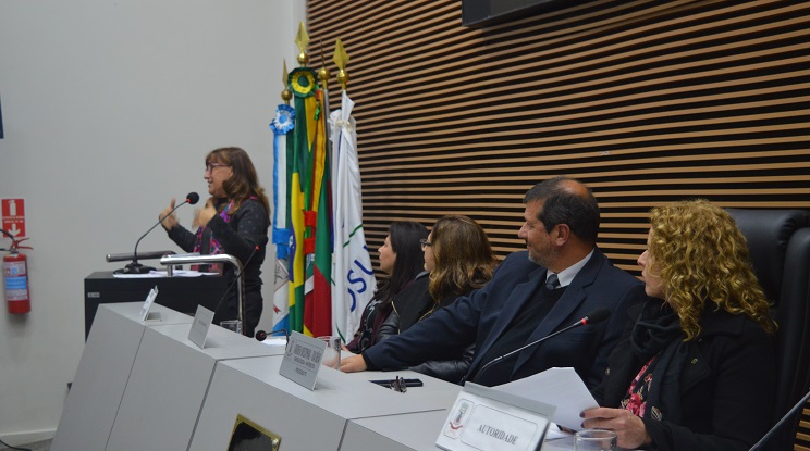 Reitora da FURG aparece discursando na tribuna e demais componentes da mesa a observam.