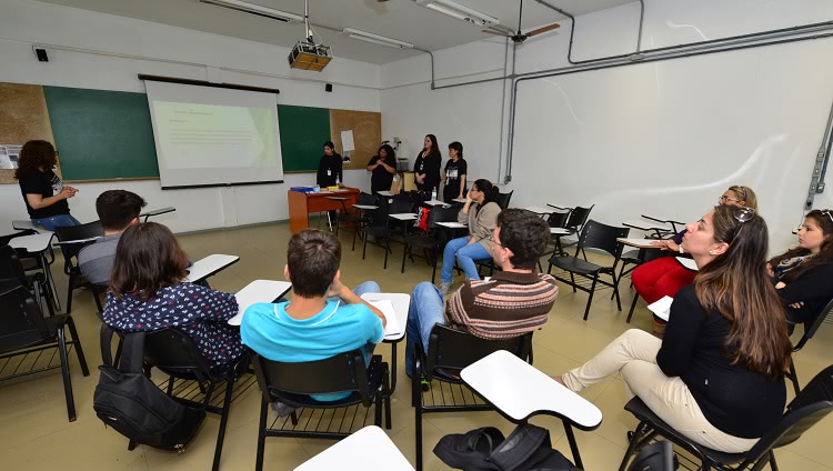 Numa sala de aula, alunos estão dispostos em meia lua, e olham para o conteúdo projetado em um telão.