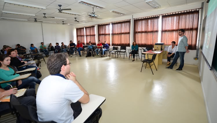 A imagem mostra uma sala de aula. Os alunos estão dispostos em círculo e olham a um rapaz que está em pé, em frente ao quadro, fazendo uma apresentação.