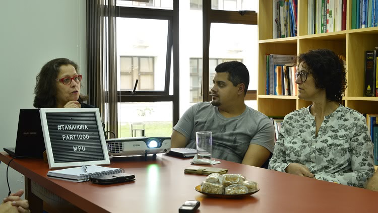 Três pessoas aparecem em um momento de reunião, no entorno de uma mesa: duas mulheres e um homem. Atrás deles, há uma janela e, ao lado, uma prateleira de livros. Sobre a mesa, um computador e uma placa, com a inscrição 