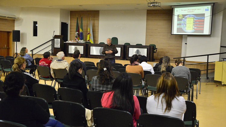 Em um auditório, o professor Cleber Palma aparece à frente do público, palestrando. 