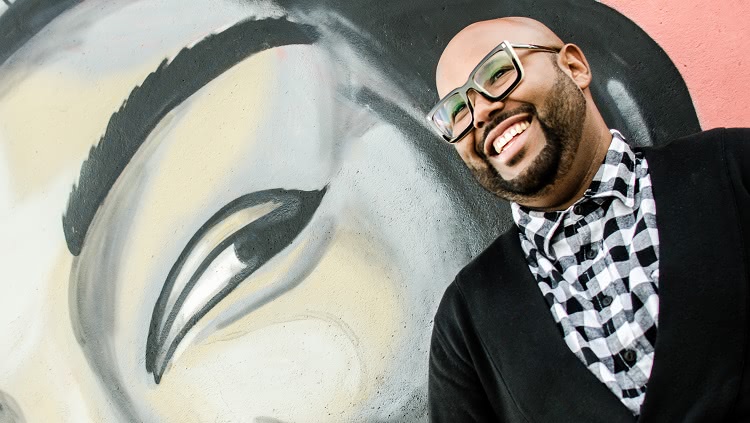 A foto foi feita na diagonal e mostra um homem negro, que usa óculos e tem barba, sorrindo. Ele aparece na parte direita da foto. Está escorado em uma parede que tem o desenho de um rosto humano em grandes proporções, do qual é possível ver o olho e a sombrancelha. 