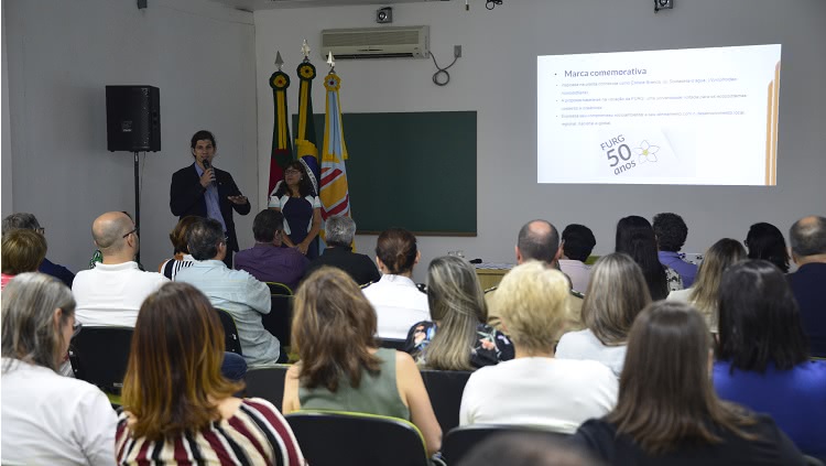 Em pé, o vice-reitor Danilo Giroldo fala ao público sentado em cadeiras no interior da sala Estuários, no Cidec-Sul. Em um telão atrás dele, há uma imagem onde se lê "Marca comemorativa" e "FURG 50 anos".