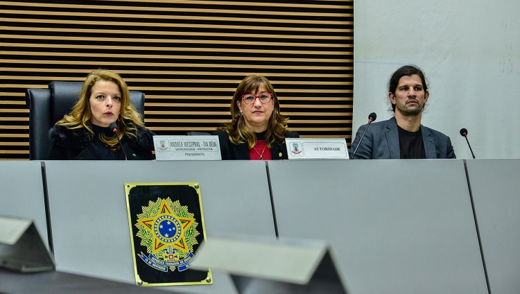 A foto é um recorte da mesa da Câmara Municipal. Aparecem três pessoas: a presidente da câmara,Tia Deia, à esquerda, ao centro, a reitora da FURG, e à direita, o vice-reitor.