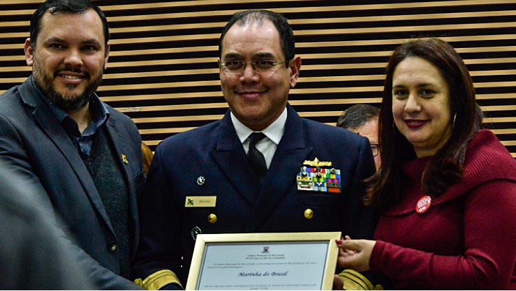 Na imagem, aparecem três pessoas. Ao centro, o vice-almirante segura um quadro. Ao lado esquerdo, um homem, ao lado direito, uma mulher. 