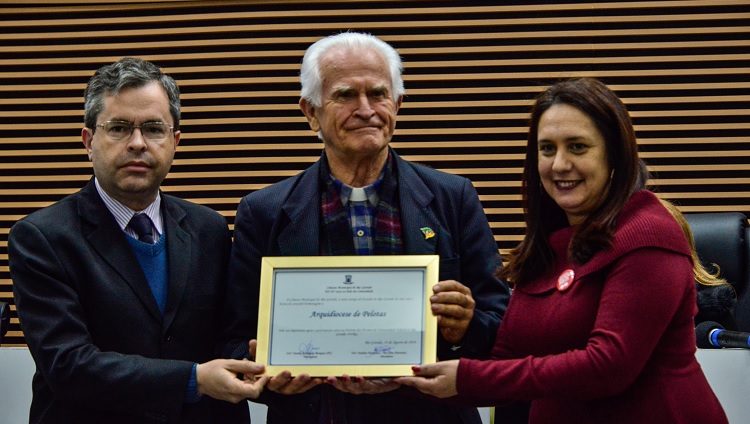 Na foto, aparece o bispo segurando um quadro. Ao lado esquerdo dele, um homem, ao lado direito, uma mulher.