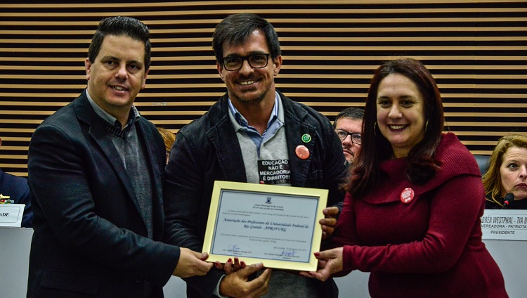 Ao centro da foto aparece o presidente da Aprofurg segurando um quadro. Ao lado direito dele, uma mulher, ao lado esquerdo, um homem. 