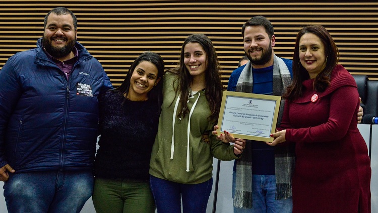 A foto mostra cinco pessoas. Duas seguram o quadro de homenagem. Todas sorriem.