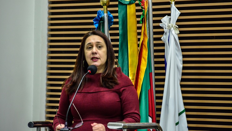 A imagem mostra em primeiro plano uma mulher, a vereadora Denise. Diante dela, há um microfone. Ela aparece da cintura para cima. Ao fundo, bandeiras do Brasil, Rio Grande do Sul e Rio Grande. 