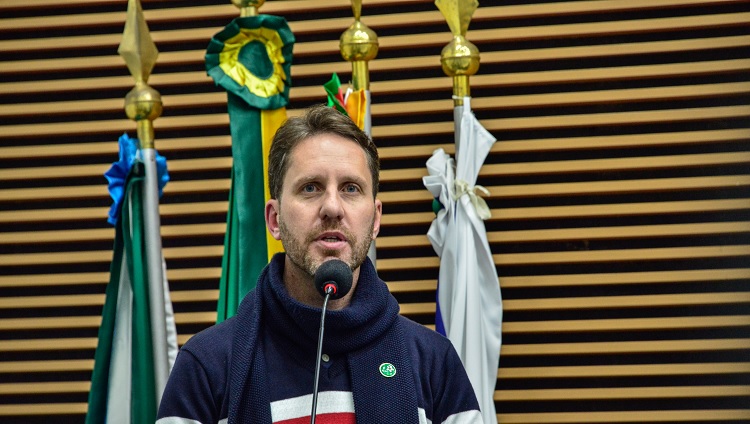 A foto mostra em primeiro plano um homem, o vereador André. O recorte é do rosto e busto. Ele fala ao microfone. Ao fundo, atrás dele, aparecem bandeiras em mastros. 