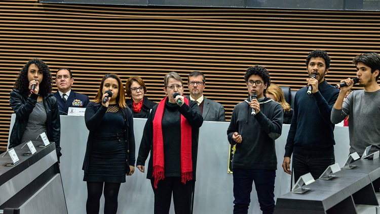 Na imagem aparecem seis pessoas cantando. À esquerda, três mulheres, à direita, três homens. Todos estão com microfones na mão. Ao fundo, aparece a mesa com as autoridades.