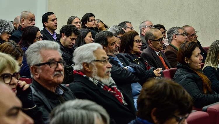 A foto mostra o público presente. As pessoas aparecem olhando em direção à mesa, que não aparece na foto. São homens e mulheres, de diversas idades. 