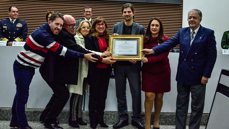 Na foto, sete pessoas, entre homens e mulheres seguram o quadro de homenagem. 