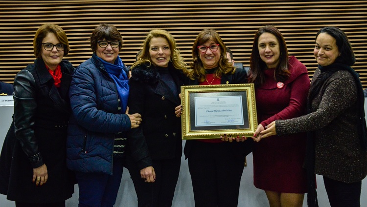 Na foto, seis mulheres posam ao lado da reitora, que segura o quadro de homenagem. 