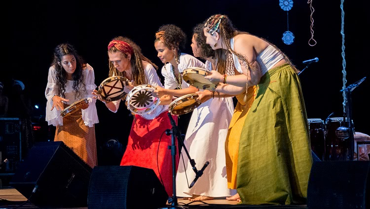 A foto mostra as cinco mulheres no palco, curvadas e tocando pandeiros. Todas estão vestindo saias longas.