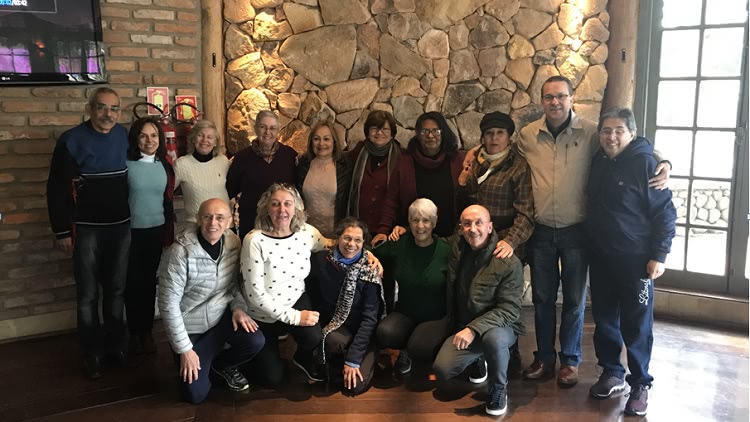 Um grupo de pessoas posa para uma foto. Há 10 pessoas em pé ao fundo, e 5 na frente, agachadas. São todas pessoas de meia idade, entre homens e mulheres. O fundo atrás deles é uma parede de pedra. À direita, também ao fundo, aparece parte de uma porta de vidro, pela qual entra claridade. 