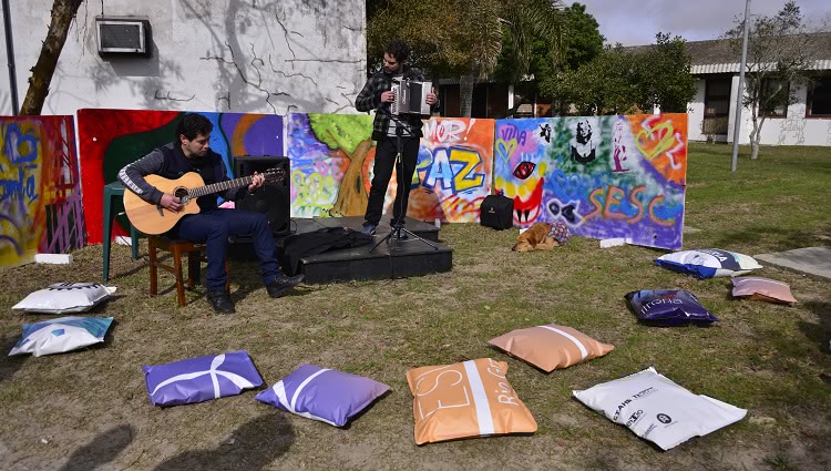 A imagem mostra duas pessoas, uma tocando violão e outra tocando uma gaita de oito baixos