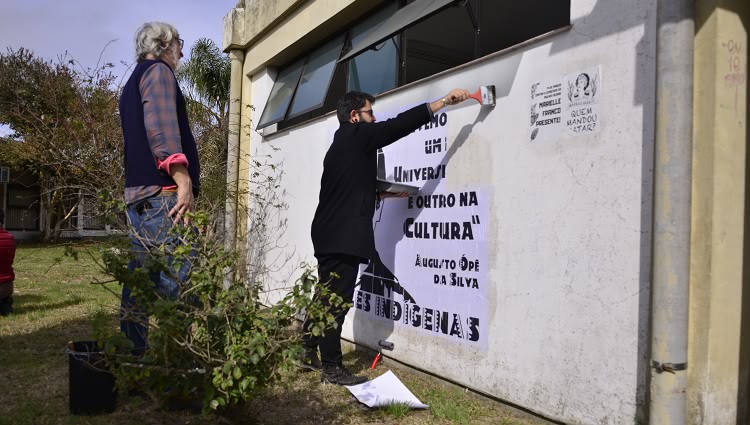 A imagem mostra duas pessoas realizando uma intervenção artística por meio de colagens em uma parede