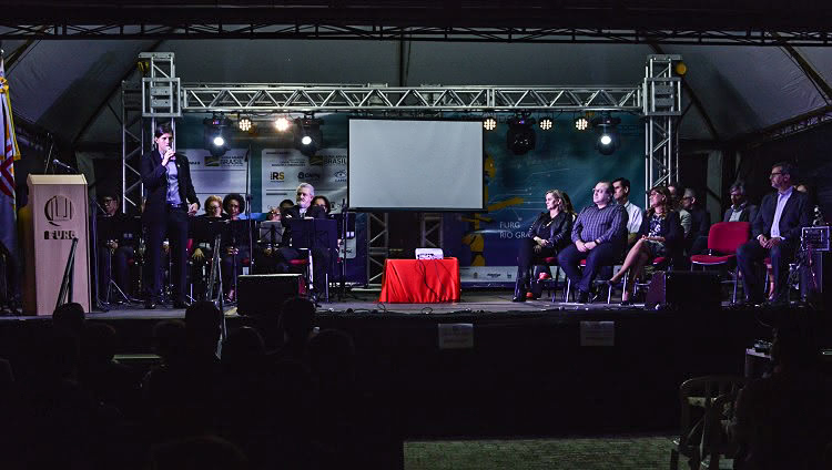 A foto foi feita à noite, num palco iluminado. Há várias pessoas sentadas em um homem em pé, com o microfone à mão