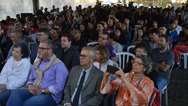 Na imagem aparece várias pessoas sentadas: é o público do evento, olhando em direção ao palco, que não aparece na foto