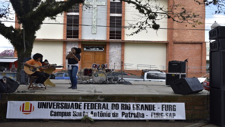 A imagem mostra uma menina em cima do palco cantando ao microfone acompanhada de um rapaz que toca violão.