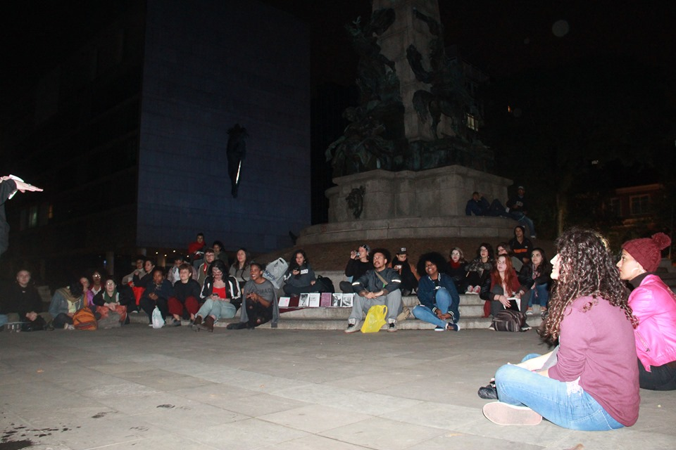 A foto foi feita à noite, numa praça. Dezenas de jovens estão sentados no chão, em formato de semi círculo, à frente de um monumento na praça.