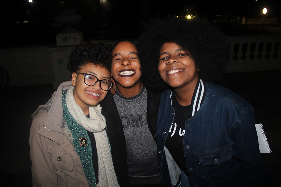 Na imagem aparecem três jovens mulheres negras. As três sorriem. 