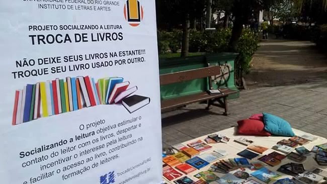 O cenário é uma praça. A imagem mostra, em primeiro plano, um banner e, ao fundo, no chão, muitos livros espalhados sobre uma manta. Há também almofadas sobre a manta. Atrás, um banco de pra.a No banner está escrito: “Projeto Socializando a leitura: troca de livros. Não deixe seus livros na estante, troque seu livro usado por outro”. 