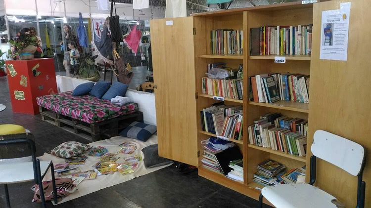 Um espaço montado na feira do livro, com um armário com as portas abertas e, em seu interior, muitos livros. Ao lado do armário há um pano no chão e, sobre ele, almofadas e livros. Ao redor do pano, uma cadeira e um banco. Próximo também há um sofá feito de pallets, com almofadas. 