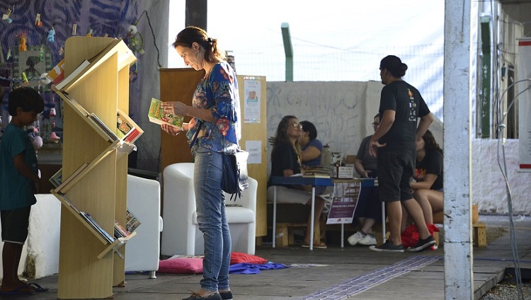 Também na feira do livro, em frente a uma estante de livros, uma mulher aparece com um livro na mão. Ela olha para o livro. Atrás, ao fundo, aparecem pessoas em pé ou sentadas, ao redor de uma mesa. 