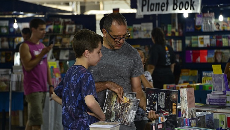 Em uma banca de livros, aparecem um homem adulto e um menino, escolhendo livros. Ao fundo, várias bancas de livros desfocadas, no espaço de vendas da Feira do Livro