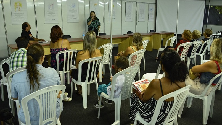 A foto mostra várias pessoas, a maior parte mulheres, sentadas, de costas para a câmera, em cadeiras, olhando para uma mulher, à sua frente, que fala atrás de uma mesa. 