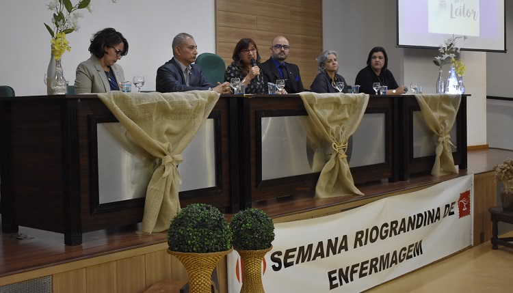 Seis palestrantes à mesa do evento. Uma deles é a reitora da FURG, no centro da mesa, e ela está falando, com o microfone à mão. Todos estão sentados. À frente da mesa há plantas e tecidos decorativos e uma faixa em que consta o texto: Semana Riograndina de Enfermagem