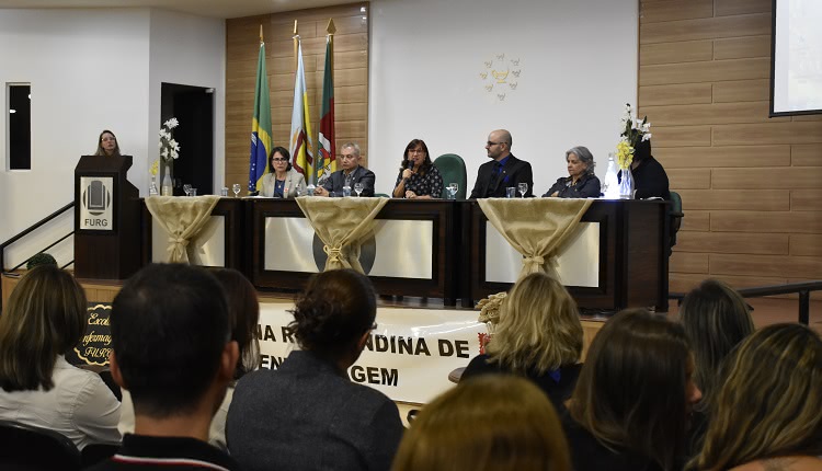 Seis palestrantes à mesa do evento. Uma deles é a reitora da FURG, no centro da mesa, e ela está falando, com o microfone à mão. Todos estão sentados. À frente da mesa há plantas e tecidos decorativos e uma faixa em que consta o texto: Semana Riograndina de Enfermagem. Nesta foto aparecem também pessoas, de costas para a foto, que estão assistindo ao evento. 