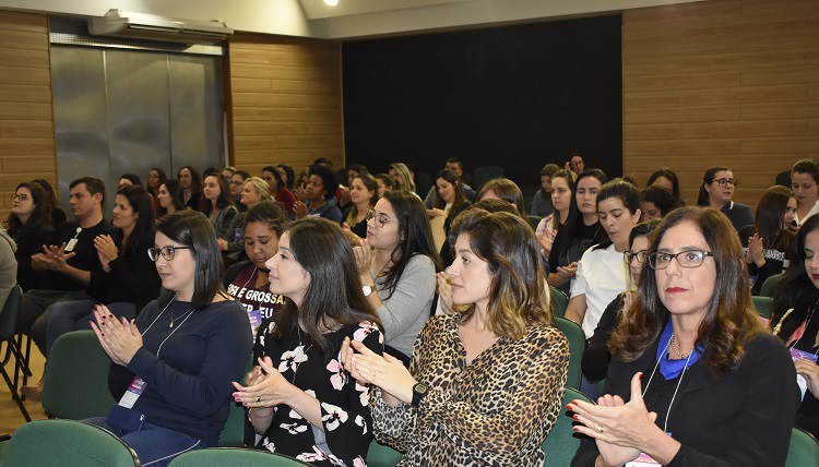 O público do evento, em um auditório aparece aplaudindo. São majoritariamente mulheres. O auditório está lotado. 