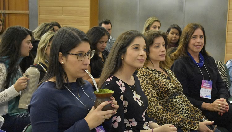 A foto mostra parte do público do evento. Uma fileira de mulheres assiste à palestra. Quatro mulheres aparecem em destaque. Uma delas toma chimarrão, as duas do lado olham para a frente, assistindo à palestra, e a última olha para a foto e sorri