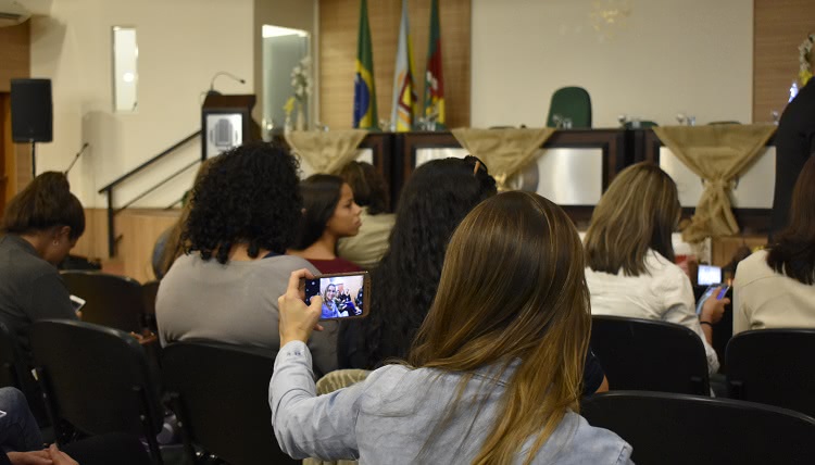 Na imagem, aparecem mulheres sentadas, de costas. À frente delas está a mesa do evento, ainda sem nenhum palestrante. Uma das mulheres está fazendo uma foto selfie com o celular. 