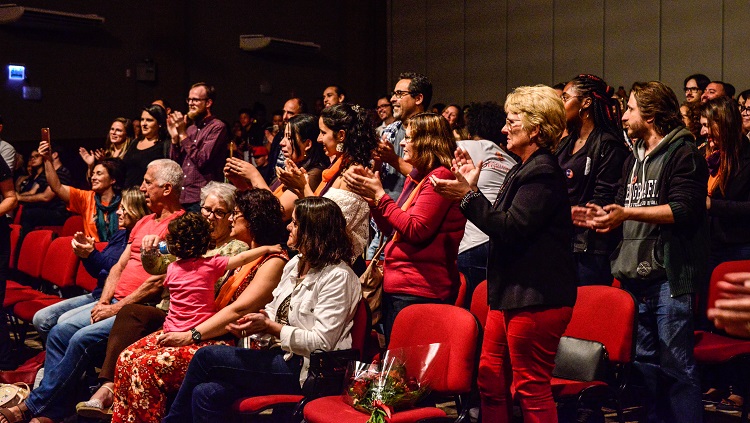 A foto mostra as pessoas da plateia, umas em pé e outras sentadas,assistindo o show