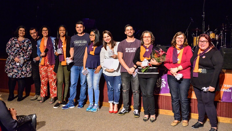 Na foto, um grupo de 11 pessoas aparecem lado a lado na frente do palco segurando as placas de homenagem entregues