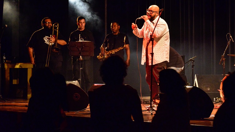 A foto mostra o vocalista Vagnotreta cantando em cima do palco e sua banca disposta ao fundo com baixa iluminação