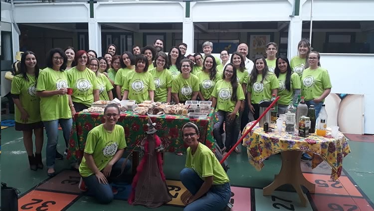 Um grupo de cerca de 30 pessoas aparece na foto, em uma cena de confraternização. A foto é pousada, todos olham para a câmera, a maior parte das pessoas atrás de uma mesa posta com lanches. À frente da mesa, duas pessoas, agachadas. No meio delas, uma boneca na forma de bruxa. Todas as pessoas usam uma camiseta verde do projeto cirandar. 