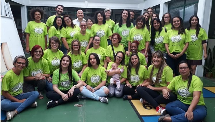 Um grupo de 32 pessoas aparece na foto, todos olham para a câmera. As pessoas ao fundo estão em pé, as mais à frente, sentadas no chão. A maior parte são mulheres, uma delas segura um bebê ao colo. Todos usam camisetas verdes do projeto cirandar. 