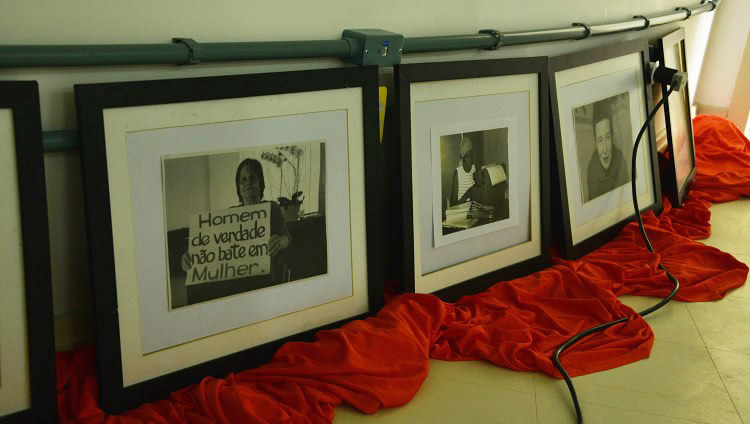 Alguns quadros com fotografias de mulheres estão colocados um ao lado do outro, apoiados na parede, em cima de um tecido vermelho. As fotografias são em preto em branco. Em uma delas, uma mulher segura um cartaz que diz "Homem de verdade não bate em mulher".