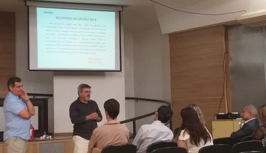 Na foto aparecem pessoas sentadas, de costas, olhando para um telão. O telão está no alto e tem como título "Relatório de gestão". Dois homens aparecem à frente do público, falando. 