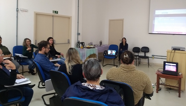 Na foto, um telão apresenta dados sobre o evento, uma cadeira com um computador e pessoas sentadas nas cadeiras